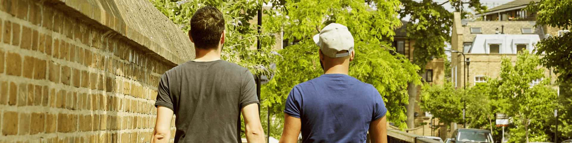 A Black person and a white person walk down a leafy, sunny street, hands held. They face away from the camera.